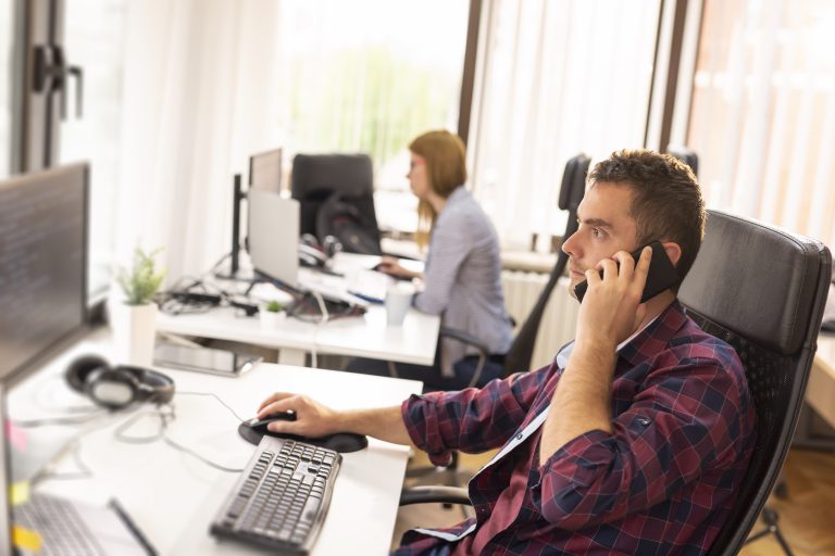 Dwóch kolegów z pracy siedzi przy biurkach w biurze, pracuje i rozmawia przez telefon; kodowanie twórców oprogramowania
