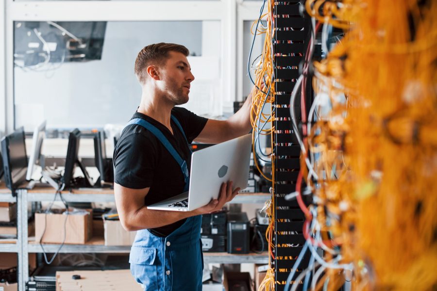 Young man in uniform and with laptop works with internet equipment and wires in server room