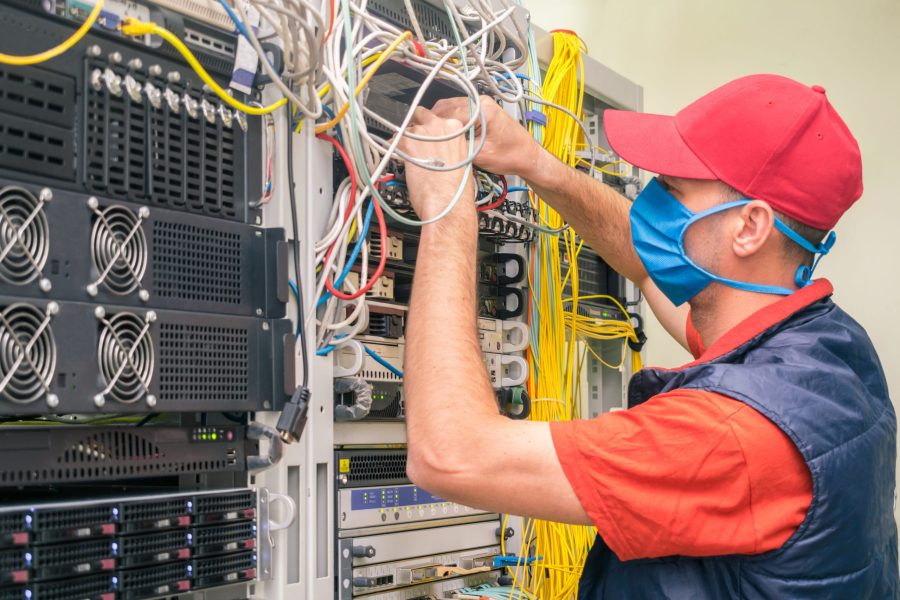 A technician in a medical mask switches wires in the server room. A man in a red cap works in a datacenter. A technical employee maintains computer equipment