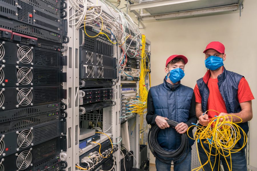 There are two technicians in medical masks in the server room. Specialists with a coil of wires in their hands work near the racks of the data center. Service engineers maintain computer equipment